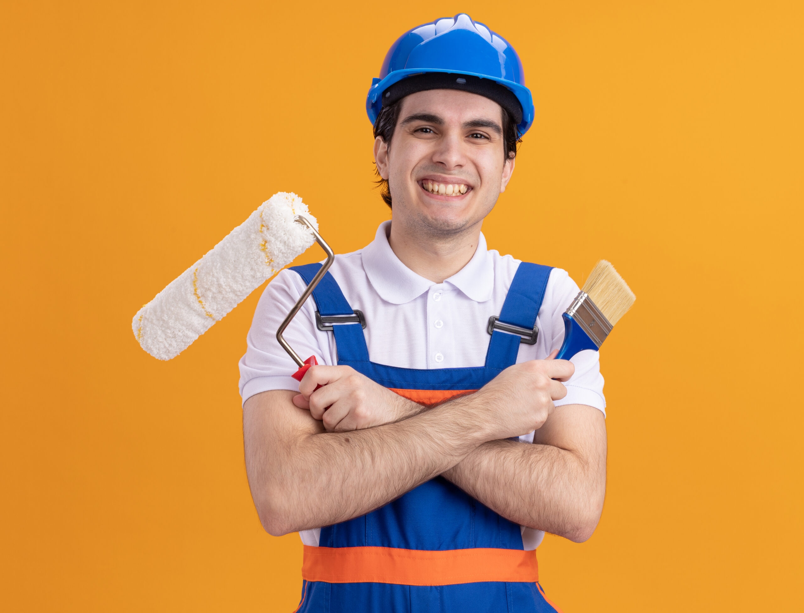 young builder man in construction uniform and safety helmet holding paint brush and roller looking at camera with big smile on face standing over orange background